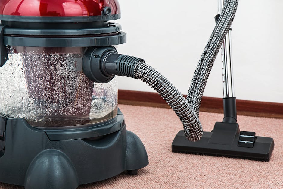 A woman with curly hair, dressed in a beige shirt and blue jeans, is kneeling on a patterned area rug in a room with warm lighting. She is inspecting a yellow and black vacuum cleaner with a flexible