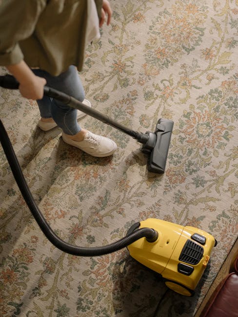 A yellow canister vacuum cleaner with a black hose and metal wand is positioned on a patterned area rug in a living room. The room features wooden flooring and a black metal media console against a wh