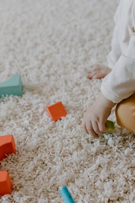 A child's hand is seen reaching out to toys on a plush, cream-colored carpet in a well-lit, domestic living room or play area. The carpet appears clean and soft, with visible fibers and a cosy texture