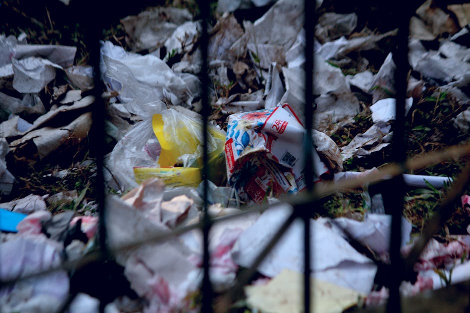 A row of crumpled black, yellow, and blue garbage bags filled with waste are piled along a white exterior wall of a building, situated on a cobblestone street. The bags contain various recyclables and