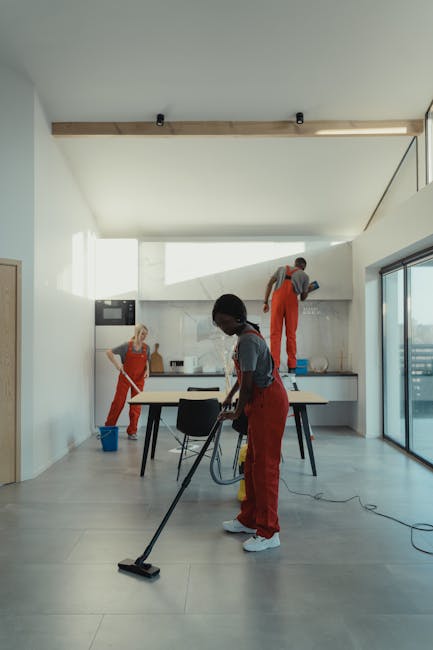 A light teal plastic dustpan with a wooden handle brush resting inside it, hanging on a small hook against a plain, neutral-colored wall. The dustpan and brush appear clean and well-maintained, indica